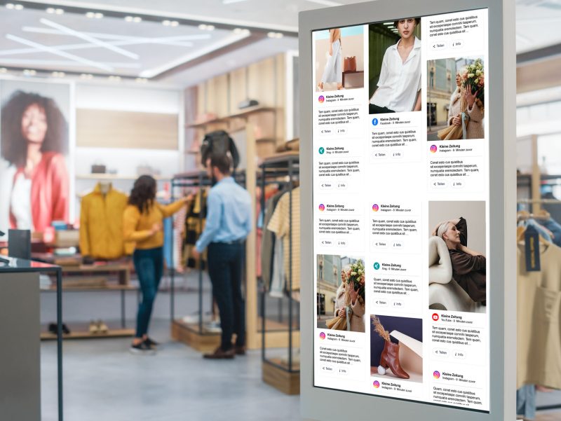 Shot of a Floor-Standing LCD Touch Screen Display with User Interface of Online Clothing Shop Standing in Clothing Store. Self service Checkout. Diverse People in Fashionable Shop Buying Clothes.