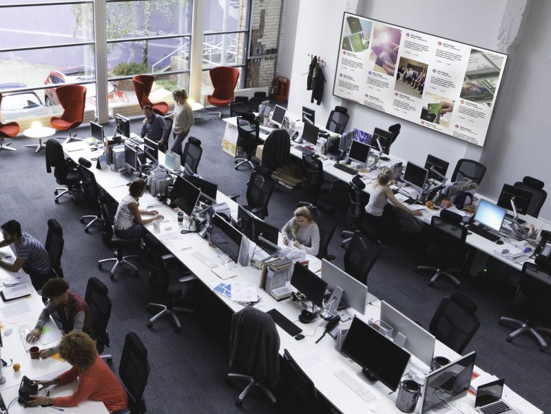 A horizontal image of a large office space shot from above. 10 office workers can be seen below. There are three large tables spread out across the room with multiple workstations.